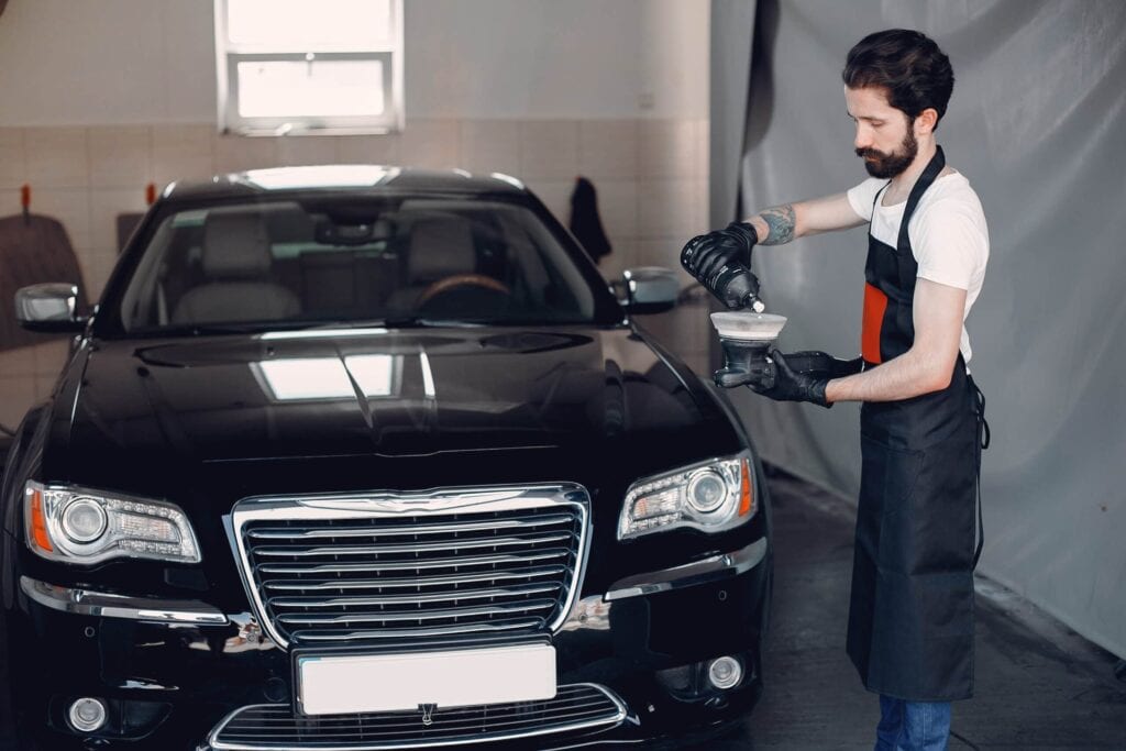 Male car detailer in a white shirt and black apron pouring a ceramic coating or polishing product from a bottle into an applicator cup while standing in front of a shiny black luxury sedan.