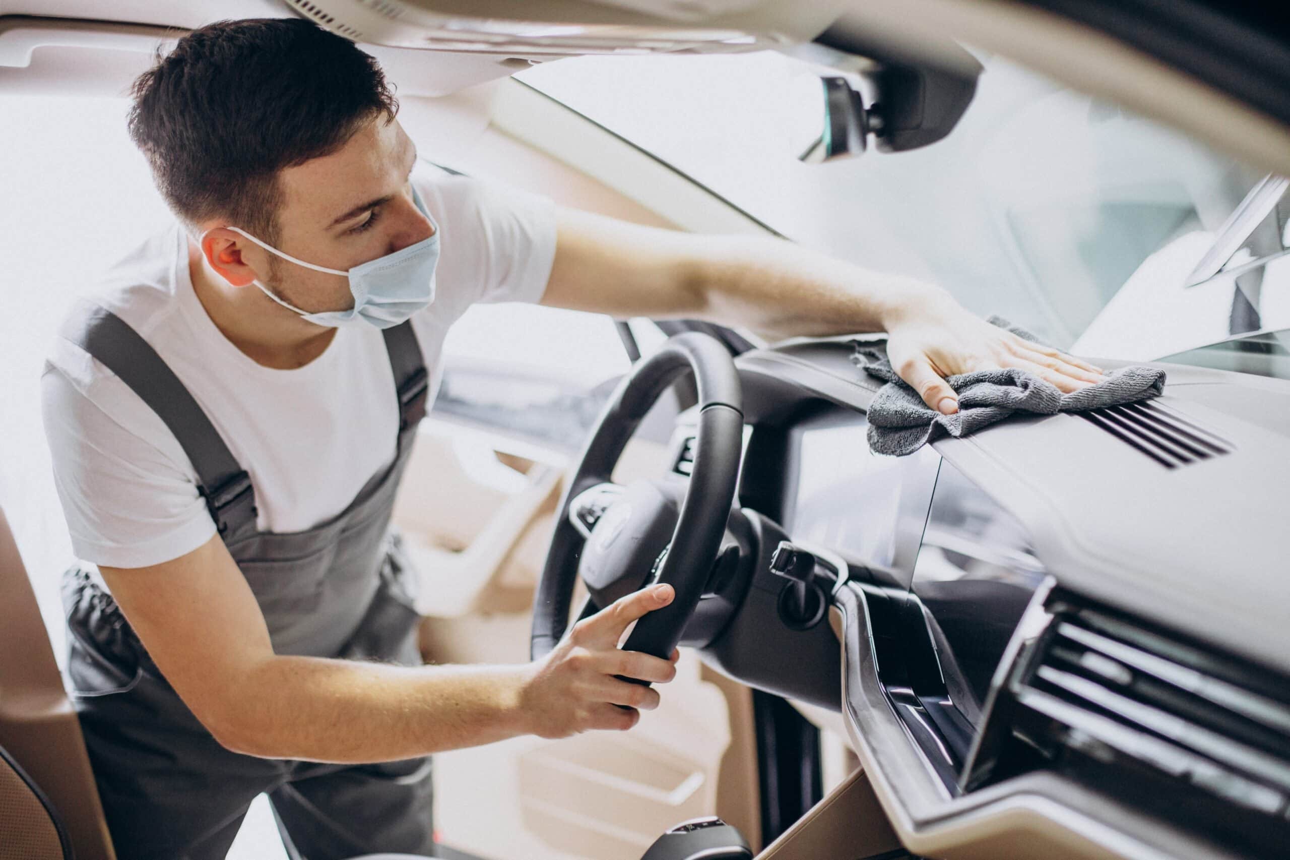 Professional car detailer wearing a face mask and grey overalls, using a microfiber cloth to wipe down and sanitize the dashboard and air vents of a modern car's interior.