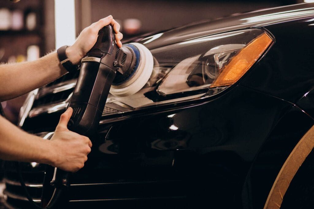 Close-up of a professional detailer's hands guiding a dual-action polisher/buffer on the glossy black hood next to the headlight of a car to remove oxidation and restore clarity.