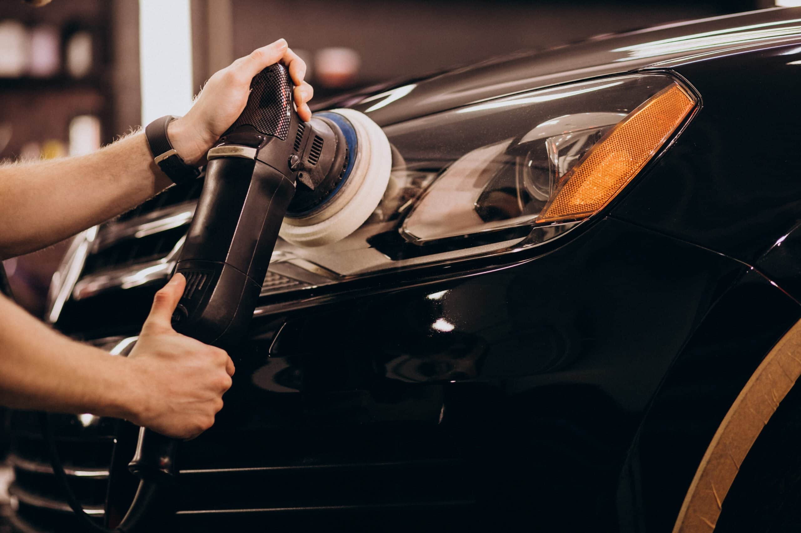 Close-up of a professional detailer's hands guiding a dual-action polisher/buffer on the glossy black hood next to the headlight of a car to remove oxidation and restore clarity.