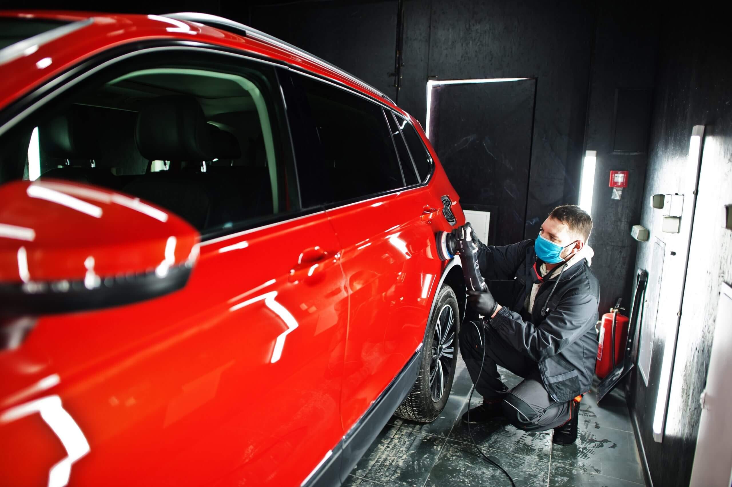 Car detailer crouching to polish the lower rear panel and wheel arch of a glossy red SUV using a dual-action buffer, highlighting the highly reflective paint finish.