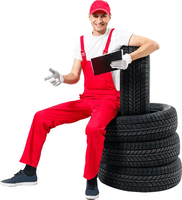 Friendly, smiling male mechanic in red overalls, white shirt, and a red cap, sitting on a stack of four new winter or all-season tires while holding a clipboard; isolated on a grey background.