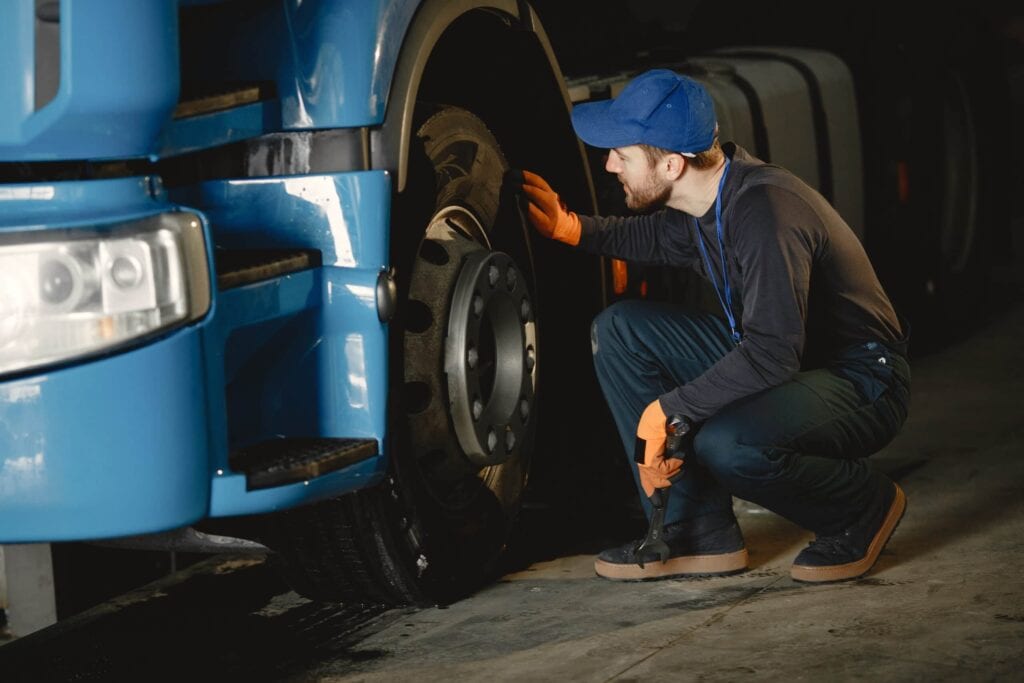 A professional mechanic in a blue cap and orange gloves inspecting a large blue semi-truck tire in a service garage.