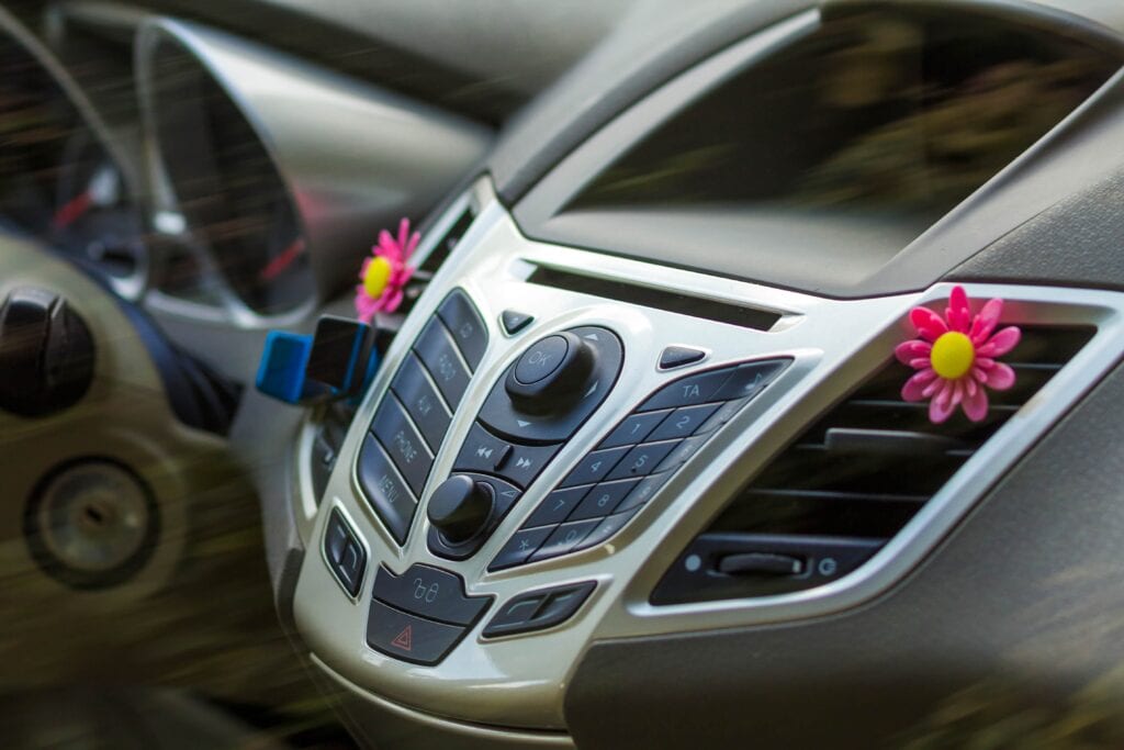 Close-up of a silver car center console with buttons for radio, phone, and menu, featuring pink flower air vent clips for interior decoration.
