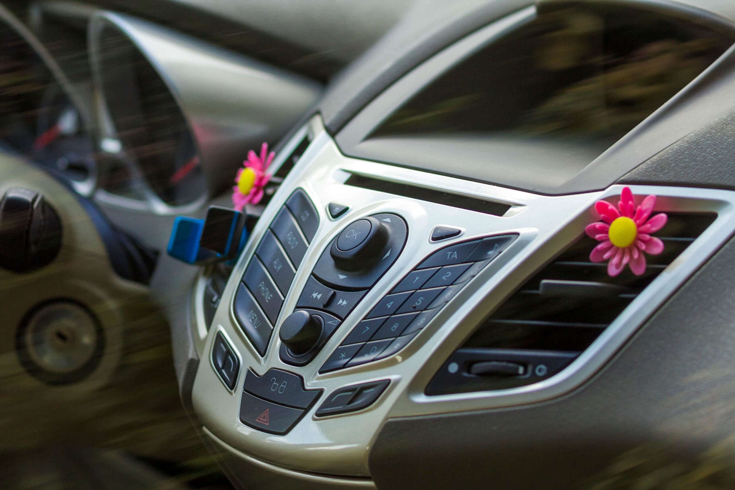 Close-up of a silver car center console with buttons for radio, phone, and menu, featuring pink flower air vent clips for interior decoration.