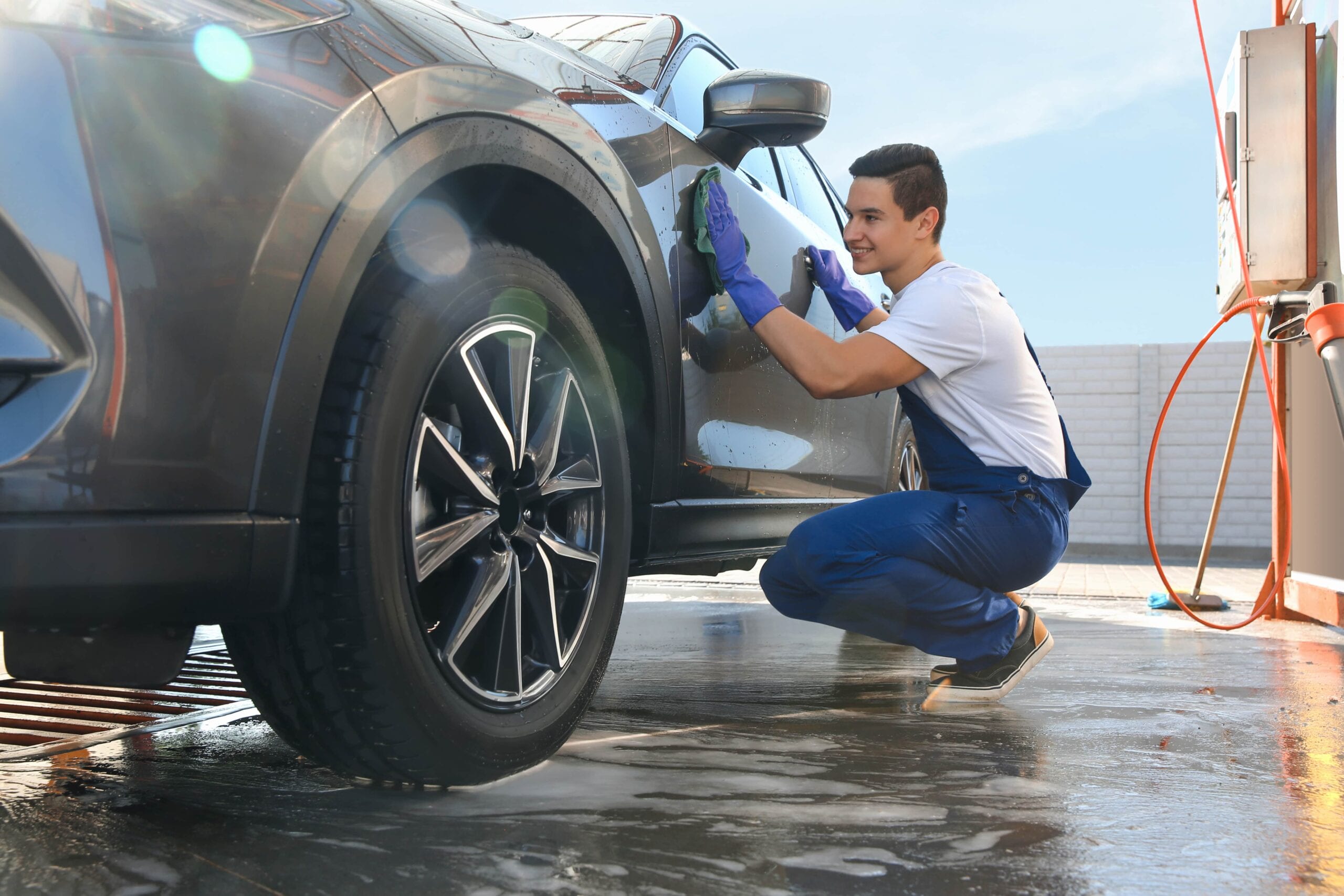 A smiling car detailing professional in blue overalls and purple gloves hand-drying a dark grey SUV with a green microfiber cloth at an outdoor wash station.