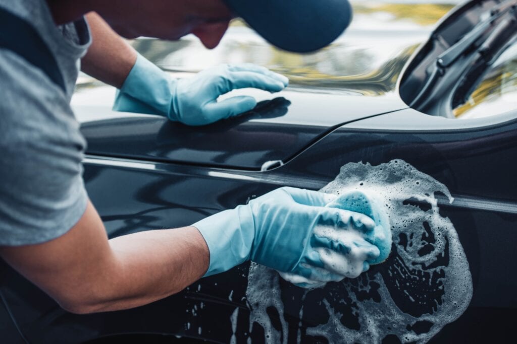 A close-up of a professional detailer wearing blue protective gloves using a soapy sponge to scrub the side panel of a dark grey car.