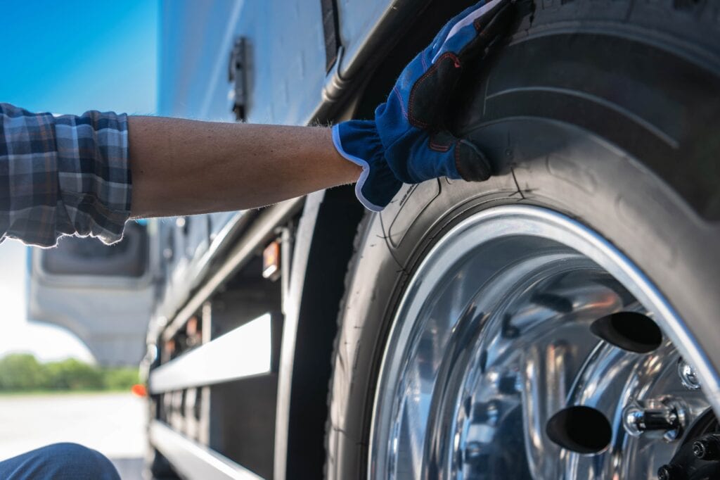 A close-up of a truck driver’s arm in a plaid shirt and work glove inspecting the tread of a large, glossy chrome-rimmed semi-truck tire.