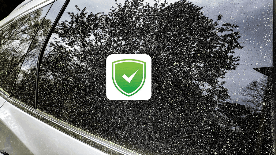 Close-up of a silver car window covered in thick yellow tree pollen featuring a green shield protection icon, offering car detailing service in Ocala, Florida.