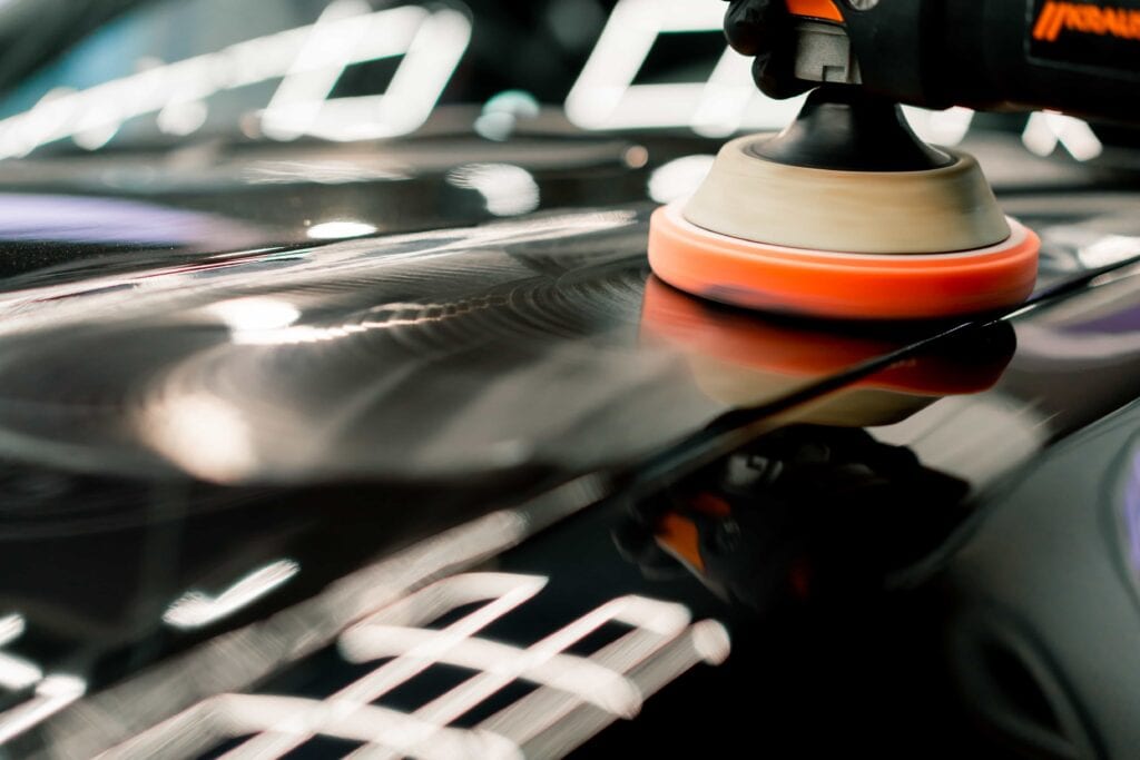 A close-up shot of an orange foam polishing pad on a dual-action orbital polisher being used on a black car hood to remove swirl marks and restore gloss.