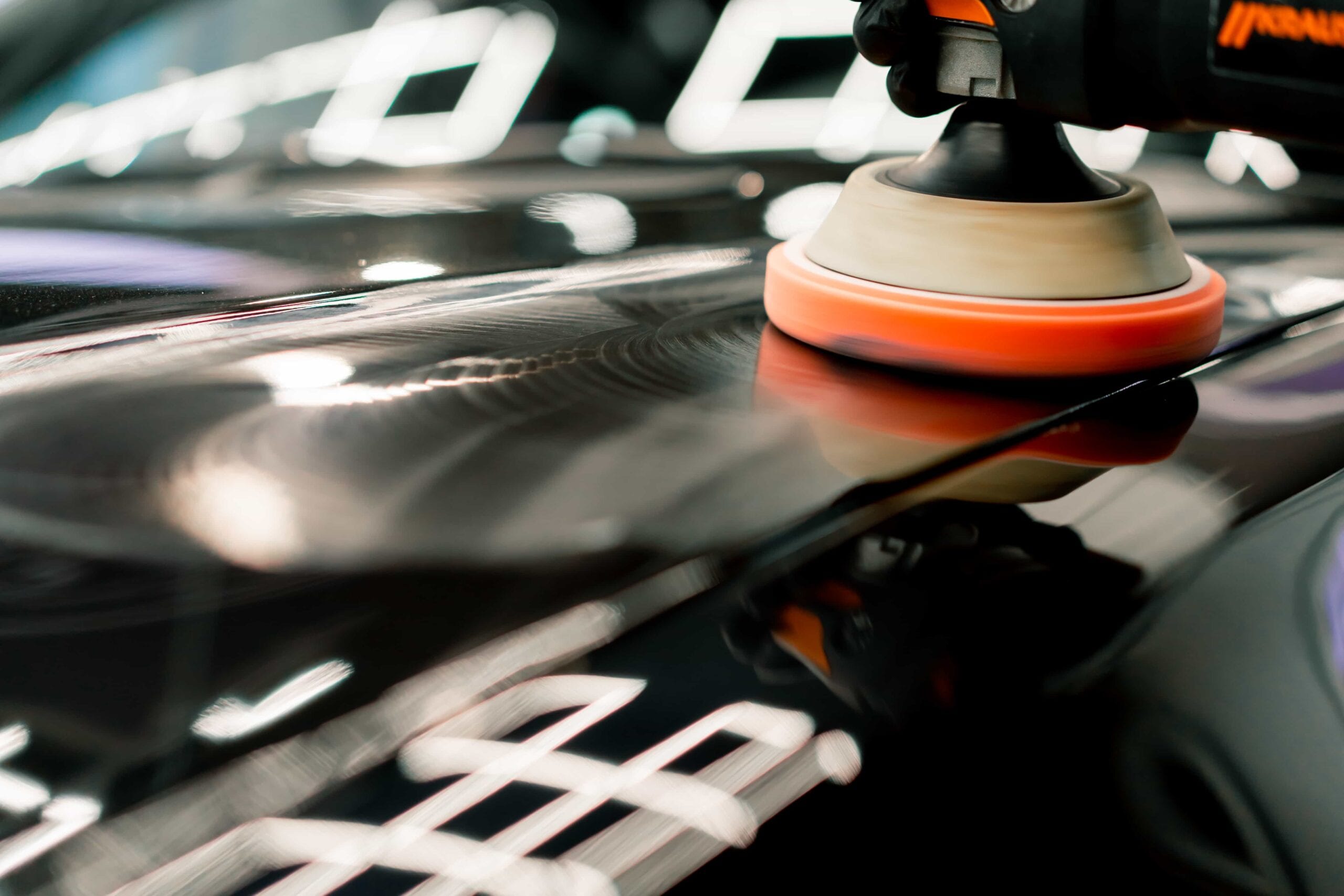 A close-up shot of an orange foam polishing pad on a dual-action orbital polisher being used on a black car hood to remove swirl marks and restore gloss.