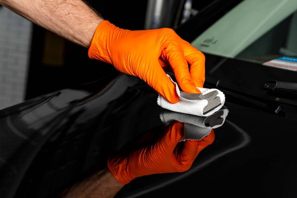 Close-up of a detailer wearing orange nitrile gloves applying a ceramic coating or wax to a black car's hood using a microfiber applicator pad.