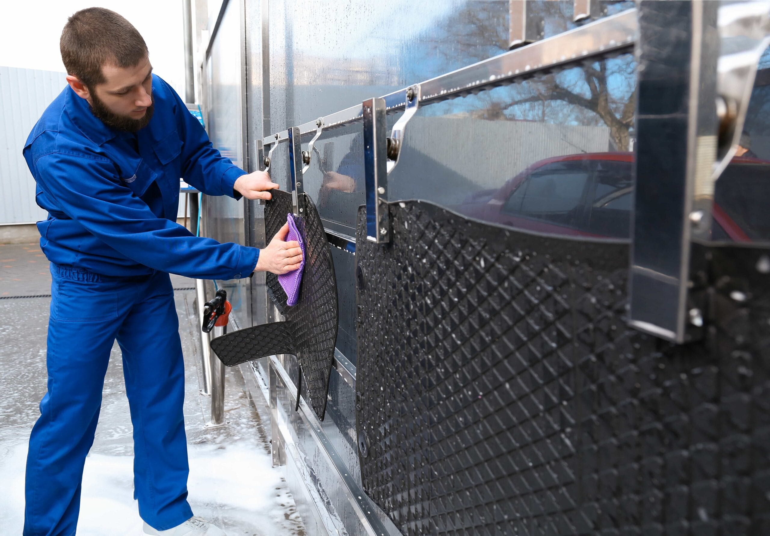 A professional car wash worker in blue coveralls uses a purple microfiber cloth to wipe down and dry black rubber car mats that are hung on a stainless steel rack.