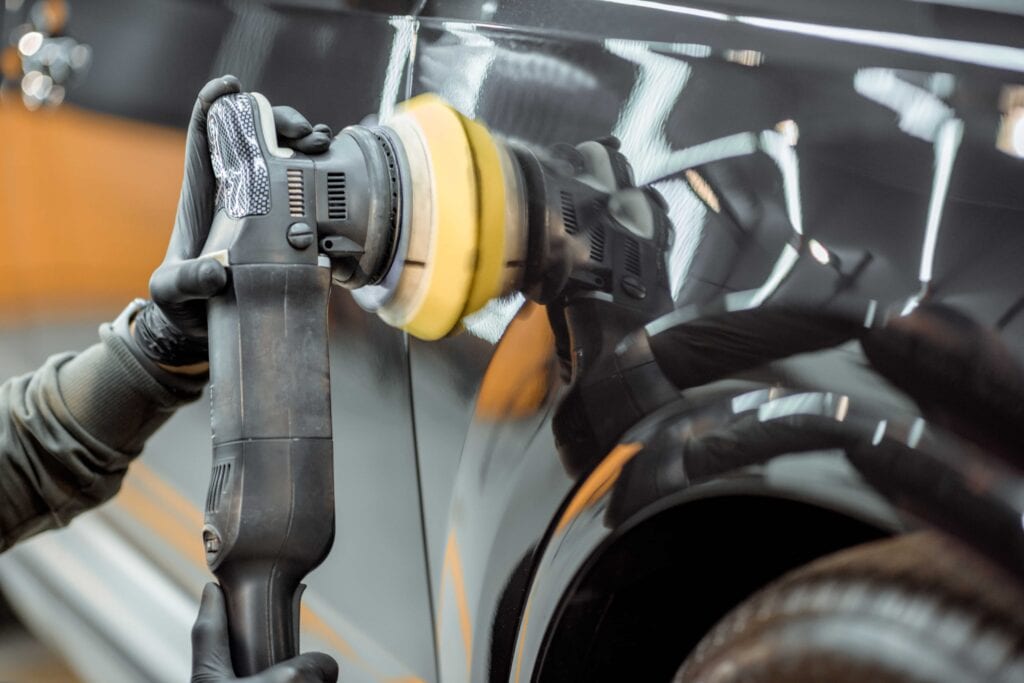 A professional detailer in black gloves using a dual-action orbital polisher with a yellow foam pad on a black car door to remove swirls and scratches.