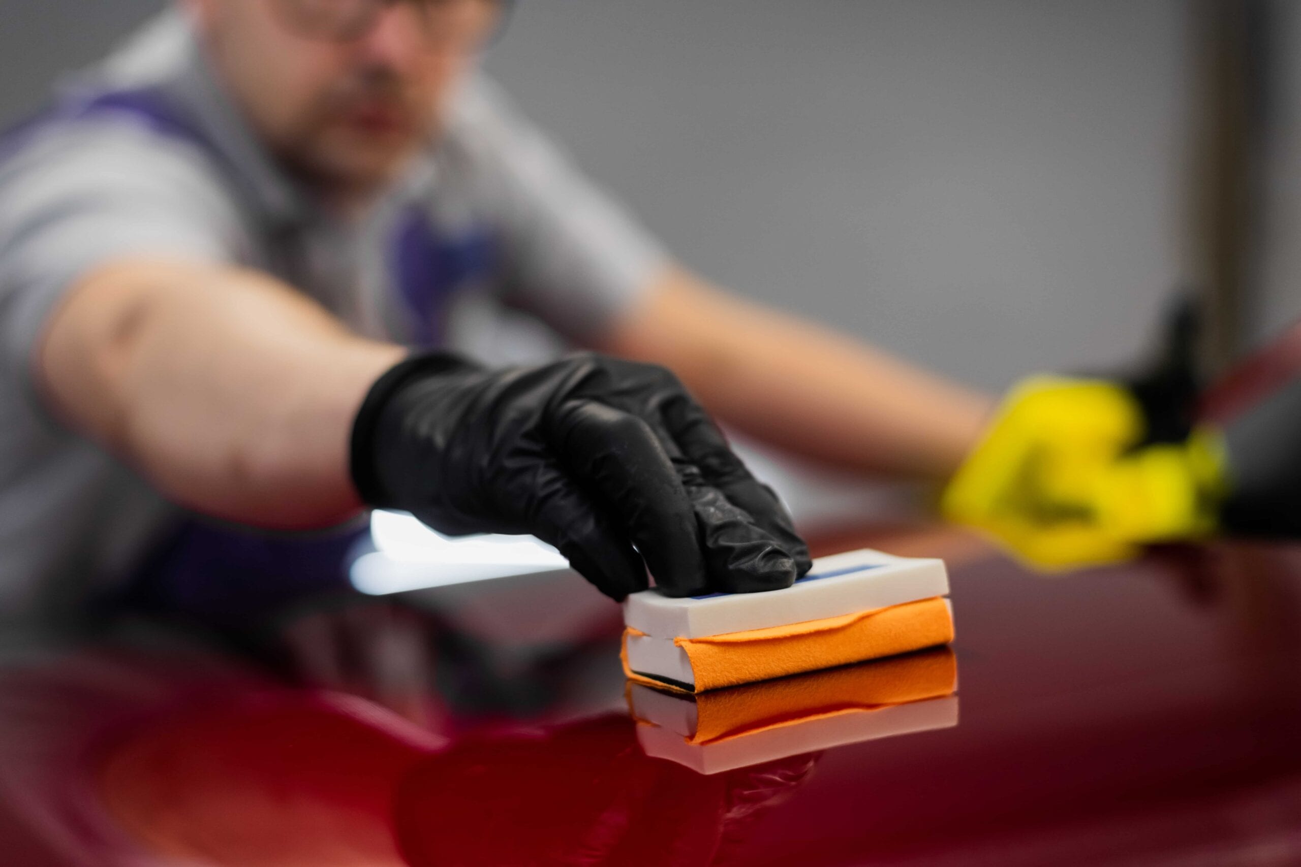 Close-up of a professional detailer in black gloves using an orange applicator block to apply ceramic coating to a glossy red car hood.