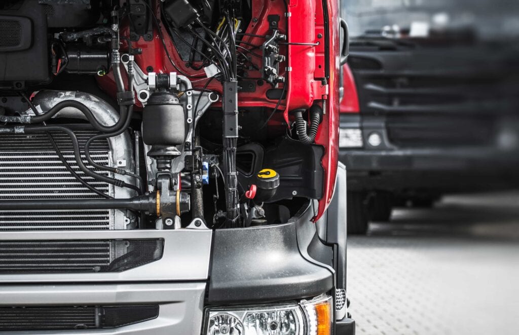 Close-up of a red semi-truck with its front hood removed, exposing the radiator, air suspension components, and complex wiring for mechanical repair at a service center.